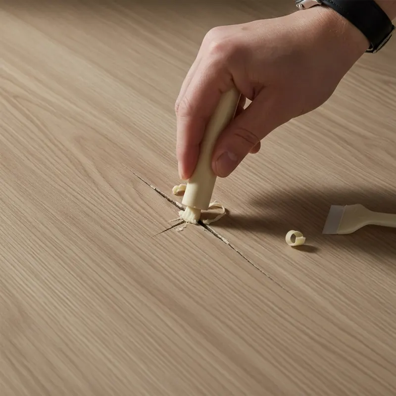 A detailed, photorealistic macro shot focusing on a person's hand carefully repairing a scratch on a light-colored laminate floor. The hand is using a color-matched wax putty stick from a repair kit to fill the scratch. The surrounding area is clean, and the lighting is bright and focused, highlighting the repair process. The mood is precise, careful, and satisfying.